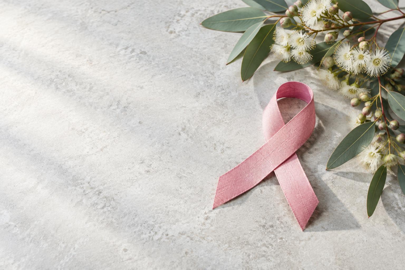 A pink awareness ribbon with eucalyptus flowers on a light stone surface